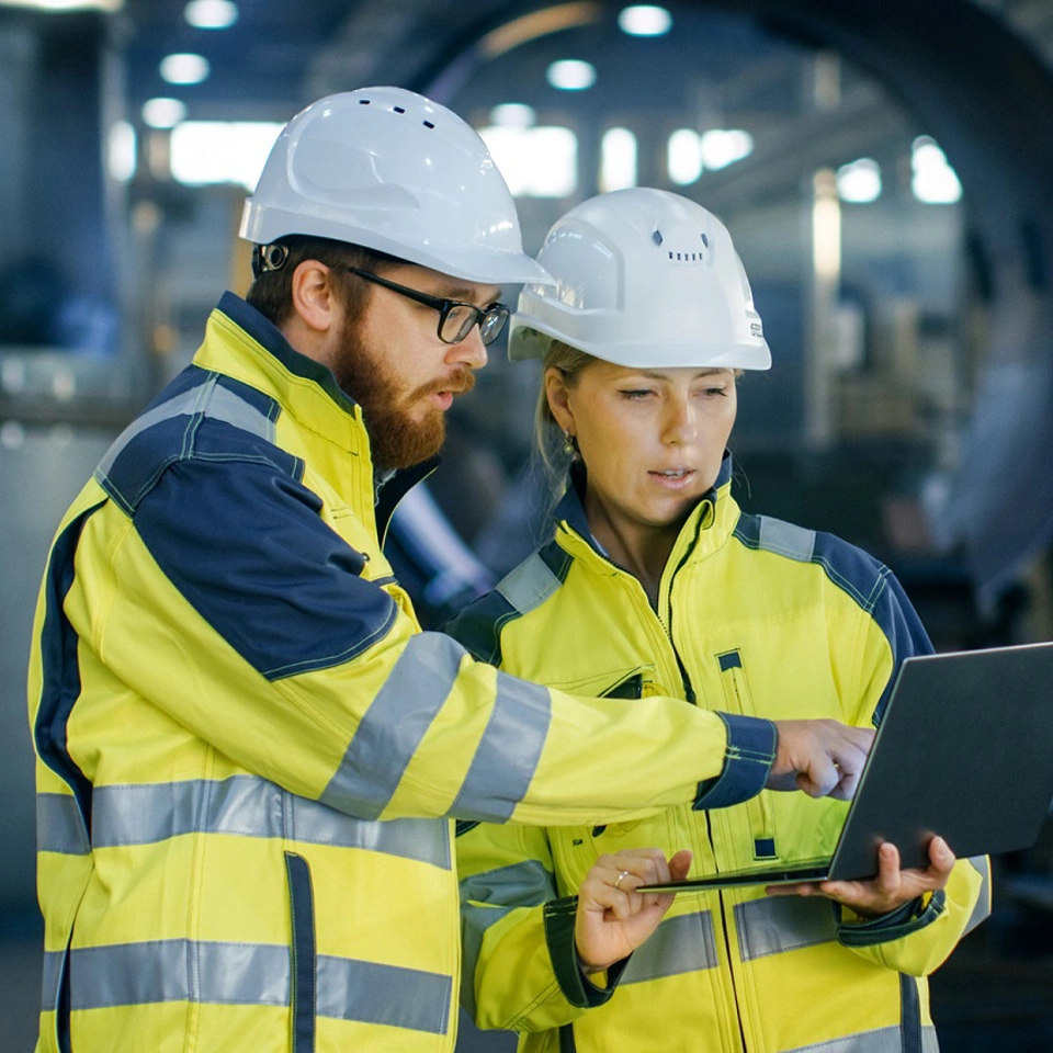 Male and Female Industrial Engineers in Hard Hats Discuss New Project while Using Laptop. They Make Showing Gestures.They Work in a Heavy Industry Manufacturing Factory.