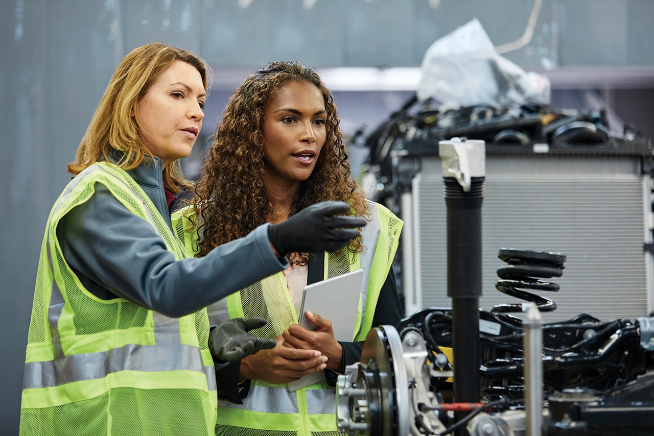 Mature female supervisor pointing to colleague by car part at factory. Confident engineers are discussing over car chassis at automobile industry. They are wearing reflective clothing.